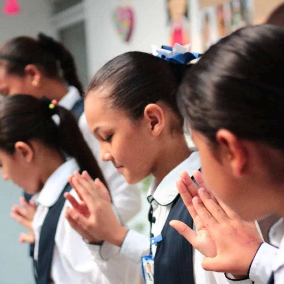 Estudiantes en uniforme escolar rezando con las manos juntas durante una actividad espiritual en el aula