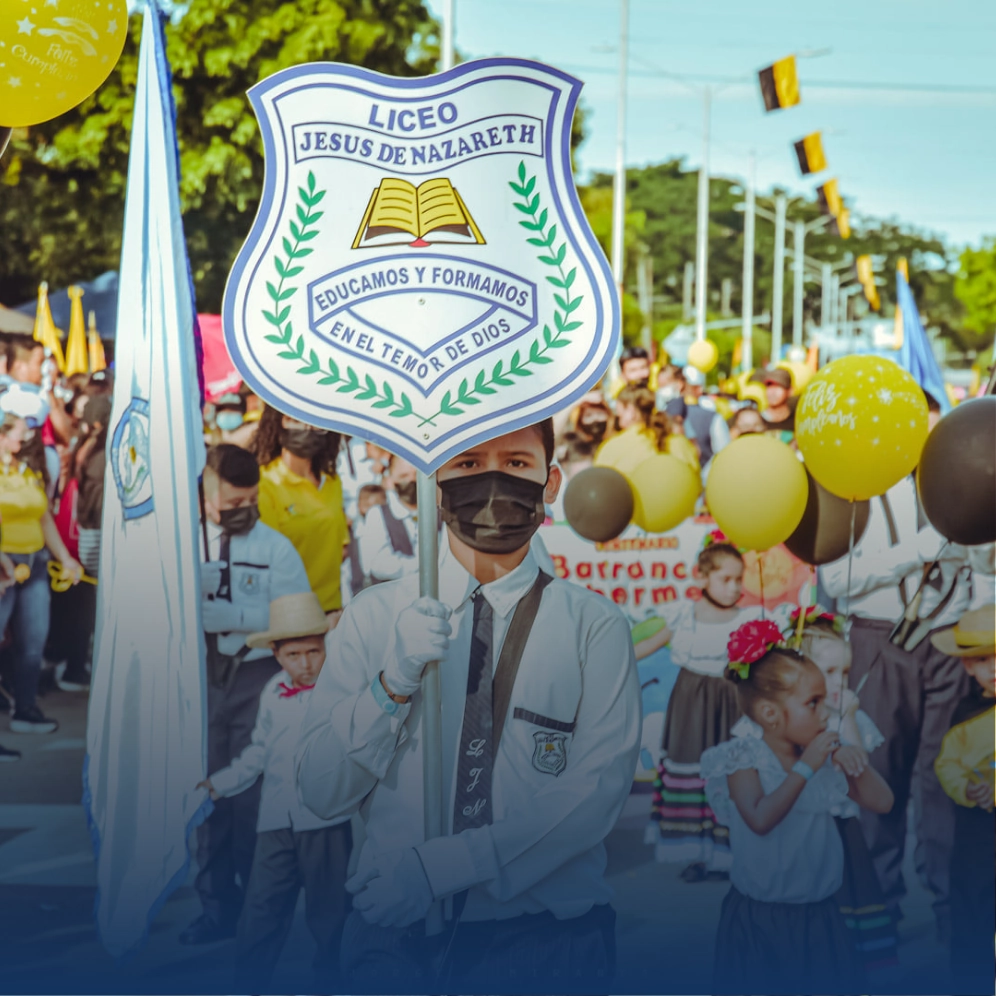 Estudiante del Liceo Jesús de Nazareth liderando un desfile institucional con el escudo del colegio al frente, acompañado por compañeros en trajes típicos y globos
